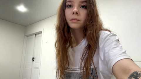 Video of a young Caucasian woman with long, wavy brown hair, wearing a white graphic t-shirt, standing in a minimalist, white-walled room with a door and ceiling light visible.