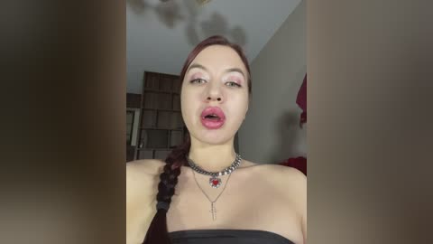 A video of a light-skinned woman with long red hair in a braid, wearing a black strapless top, silver necklace, and red lipstick. She's making a pouty face, with a blurred background of wooden shelves and a red garment hanging.