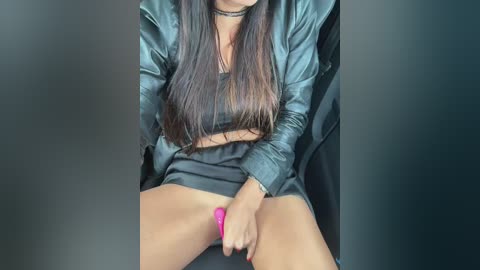 Video of a woman with long dark hair, wearing a black leather jacket and skirt, sitting with legs apart, holding a pink vibrator against her shaved vulva.