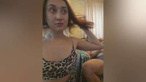 Video of a young woman with light skin and straight brown hair, wearing a leopard-print dress, sitting on a bed in a dimly lit room.