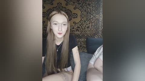 Video of a young, fair-skinned woman with long, straight, light brown hair, wearing a black t-shirt and a necklace, sitting on a bed with patterned wallpaper in the background.