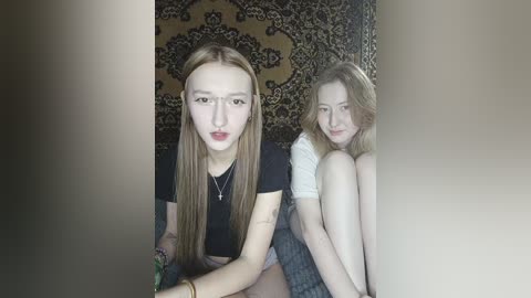 Video of two young women with fair skin, long hair, and similar facial features. They sit back-to-back, wearing casual outfits. Background features an ornate, dark-patterned rug.