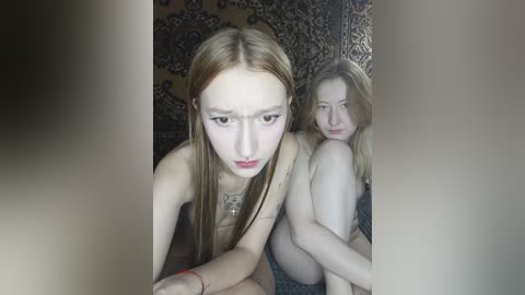 Video of two young women with pale skin, long straight hair, and similar facial features, one topless, the other partially clothed, sitting on a patterned rug.