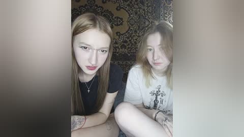 Video of two young women with pale skin and long, straight hair. One wears a black t-shirt, the other a white t-shirt. They sit side by side on a patterned rug, both looking slightly bored.