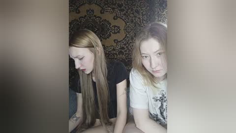 Video of two young women with long hair, one in a black shirt, the other in a white graphic tee, standing close in a dimly lit room with patterned wallpaper.