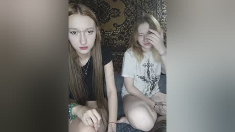 Video of two young women with pale skin and straight brown hair, wearing casual outfits; one in a black top and bracelet, the other in a white graphic tee, sitting on a dark couch in a dimly lit room with patterned tapestry backdrop.