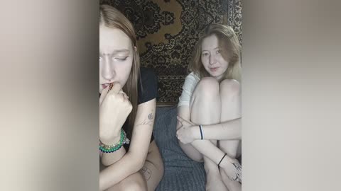 A video of two young women in a dimly lit room, one in a black top, the other in a white top, sitting on a bed, with a tapestry and a patterned rug in the background.