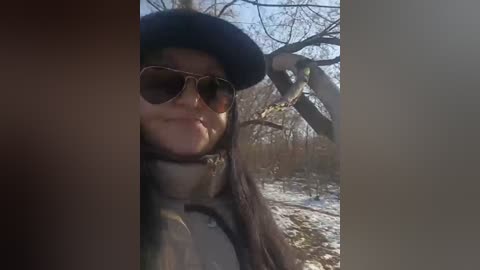 Video of a woman with light brown skin, wearing a dark blue beret, aviator sunglasses, and a beige puffer jacket, standing in a snowy, leafless forest, with sunlight filtering through the branches.