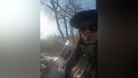 A video of a woman with long brown hair, wearing sunglasses and a black hat, holding a can of beer, standing in a forest with leafless trees and sunlight filtering through.