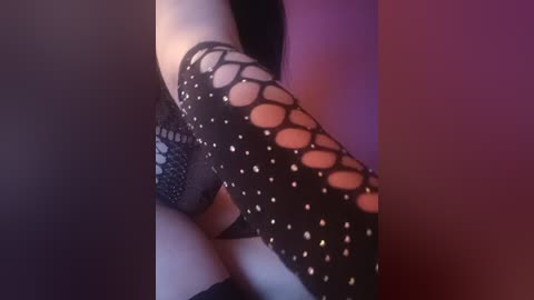 Video of a woman's arm in black fishnet stockings with a diamond pattern, set against a purple background. Her skin is pale, and her hand is partially visible. The image is close-up and slightly blurred.