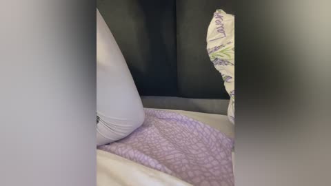 A video showing a person in white pajamas lying on a bed, covered with a light purple quilt and a patterned pillow, with a black headboard in the background.