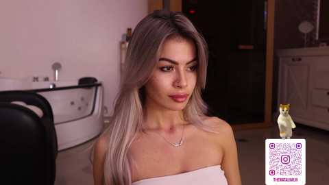 Video of a young woman with long, platinum blonde hair, wearing a strapless, white top, in a modern bathroom with a white bathtub, sink, and QR code watermark.