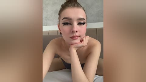 Video of a young Caucasian woman with light skin, brown hair in a bun, and a strapless dark top, leaning on her elbows, looking directly at the camera, in a minimalist room with a gray wall.