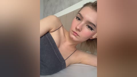 Video of a young Caucasian woman with light skin and long blonde hair, wearing a gray ribbed tank top, lying on a bed. She has a pensive expression, with prominent eyeliner and makeup.
