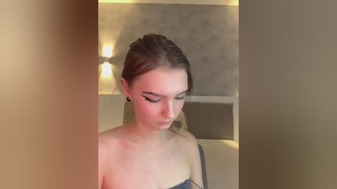 A video of a young, light-skinned woman with long, straight, brown hair tied back, wearing black eyeliner, standing in a dimly lit, beige-walled room with a tufted headboard in the background.