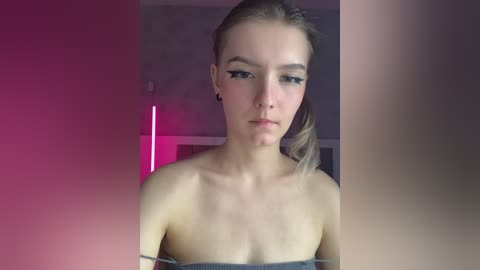 Video of a young, fair-skinned woman with blonde hair tied back, wearing a strapless top, standing indoors against a muted, gradient background.