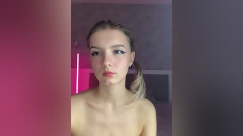 Video of a young, fair-skinned, topless woman with blonde hair in a ponytail, looking serious. Background features a dimly lit, minimalist room with a pink and purple gradient.