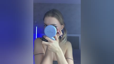 Video of a young, light-skinned woman with blonde hair, partially nude, holding a compact mirror, applying makeup in a dimly lit, minimalist bathroom.