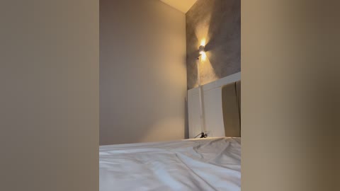 A video of a minimalist bedroom with a beige wall and a white bed, featuring a small, modern lamp emitting a warm glow. The room is sparsely decorated, emphasizing simplicity and clean lines.