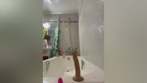 A video of a modern bathroom featuring a white sink with a brown dildo and a green shower curtain. The room is tiled in beige, with visible toiletries on the counter. A person, partially visible, is using the sink.