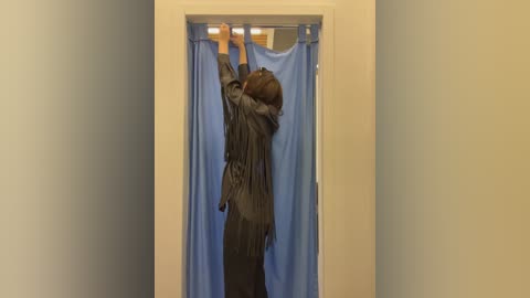 A video of a woman with shoulder-length brown hair, wearing a dark grey pinstripe suit, adjusting a blue curtain in a beige hallway.