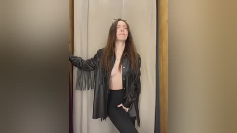 Video of a young woman with long brown hair, wearing a black leather jacket, black pants, and no top, leaning against a beige curtain in a dimly lit room.