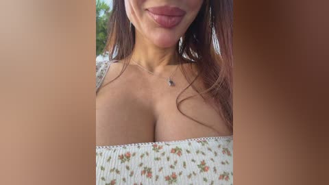 Video of a smiling woman with long brown hair, wearing a floral off-shoulder top, showing cleavage, with a necklace, against a blurred outdoor background.