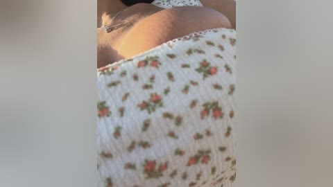 A close-up video of a person's upper body, showing a portion of their back and shoulder. They wear a white, floral-patterned dress with green and red flowers, and a silver necklace. The background is blurred, emphasizing the subject.