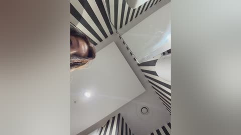Video of a person with a shaved head and a beard, standing in a room with white walls and black and white striped wallpaper, viewed from a low angle.