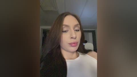 Video of a young woman with long brown hair, fair skin, and a white tank top, looking contemplative indoors, blurred background.