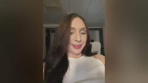 Video of a smiling Asian woman with long black hair, wearing a white sleeveless top, in an office setting with white ceiling tiles and a desk.