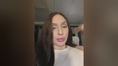 Video of a light-skinned woman with long, straight dark hair, wearing a white top, standing indoors, slightly out of focus, with blurred background of a wardrobe and ceiling.