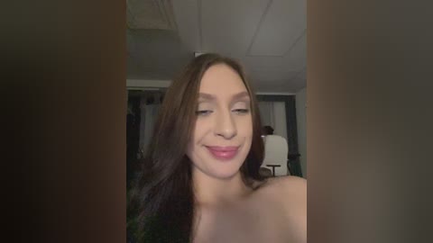 Video of a smiling, fair-skinned woman with long, dark brown hair, standing topless in a dimly lit room with a white ceiling and a white chair in the background.