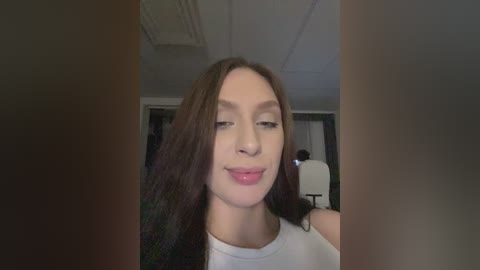 Video of a young woman with long brown hair, fair skin, and a slight smile, wearing a white tank top, taken indoors with a blurred background.