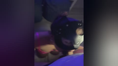 Video of a young woman in a black latex mask, kneeling with her head down, possibly in a BDSM scene, dimly lit room.