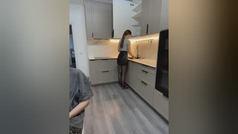 Video of a modern kitchen with a woman in a gray dress and black stockings, washing dishes; wooden floor, white cabinets, and a spiral staircase in the background.
