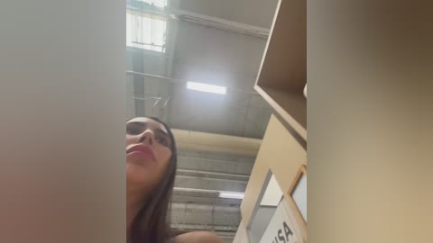 Video of a young woman with long brown hair, wearing red lipstick, looking upwards in an industrial space with beige walls and metal beams.