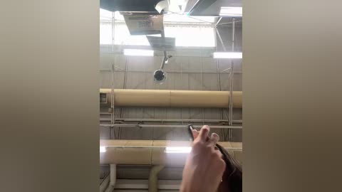 Video of an industrial room with beige walls, fluorescent lights, metal pipes, and a person holding a phone, partially visible.