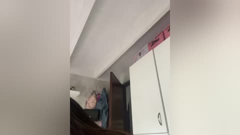 A video of a messy, dimly lit room with a person sleeping on a bed covered in clothes, under a white ceiling with a red light fixture.