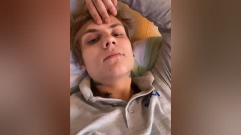 A video of a young man with fair skin, short blonde hair, wearing a grey hoodie, lying on a bed, with a hand resting on his forehead.