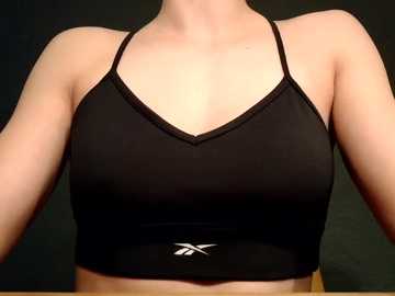 Video of a woman's upper torso, wearing a black Nike sports bra with thin straps and a small logo at the bottom. She has fair skin and is standing against a dark, blurred background.