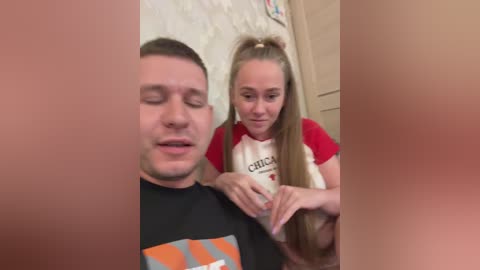 Video of a young man with short brown hair wearing a black T-shirt and a woman with long blonde hair in a high ponytail, both looking at a smartphone screen.