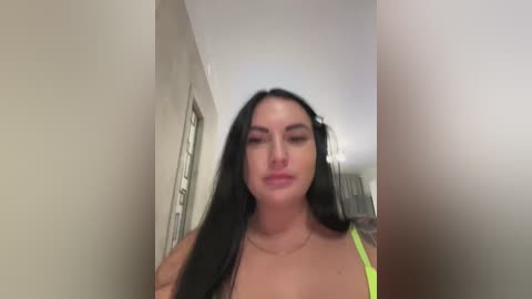 Video of a young woman with long black hair, fair skin, and a small tattoo on her left arm, wearing a neon green bra, standing indoors in a dimly lit hallway.