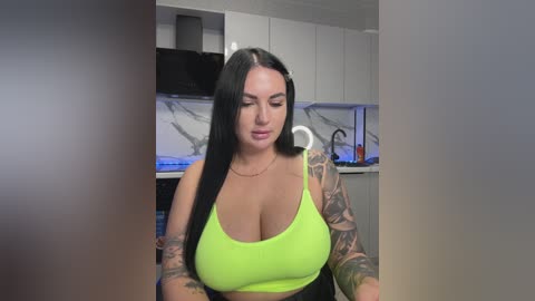 Video of a tattooed woman with long black hair, wearing a neon green crop top, sitting in a modern kitchen with white cabinets and a black stove.