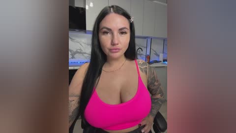 Video of a tattooed woman with long black hair, wearing a bright pink sports bra, sitting in a modern kitchen.