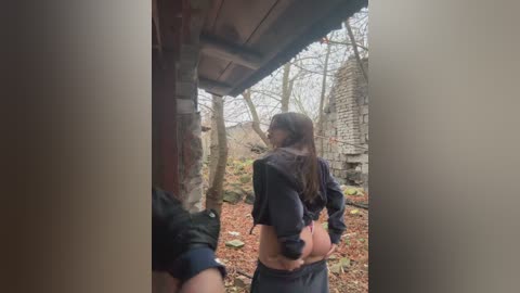 Video of a young woman with long brown hair, wearing a black crop top and low-rise jeans, standing in a dilapidated, leaf-strewn yard with a ruined building in the background.