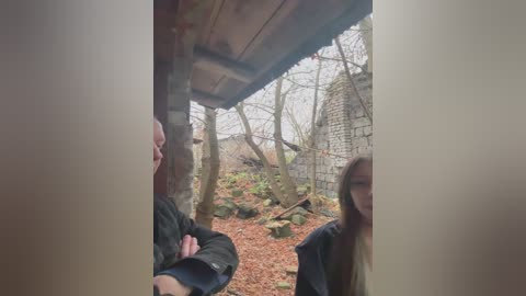 A video captures three people in a rustic, outdoor setting with bare trees, a wooden hut, and a brick wall. The ground is covered in autumn leaves.