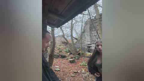 Video of a man and a woman standing near a wooden hut in a forested area with fallen leaves, partially obscured by vertical white lines.