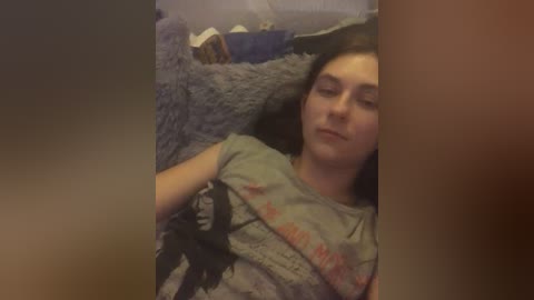 A video of a young woman with light skin and long brown hair lying on a grey, textured blanket. She wears a grey t-shirt with a black graphic. The background is dimly lit, with a soft, blurred texture.