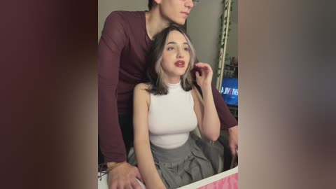 A video captures a young woman with medium-length, dark hair and fair skin, wearing a white sleeveless top and gray skirt. She sits indoors, with a man in a burgundy shirt behind her.